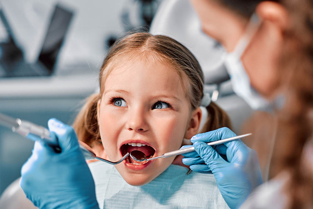 Children's dentistry. First examination at the dentist. A cute beautiful girl with an open mouth is looking to the side while the doctor is treating her teeth.
