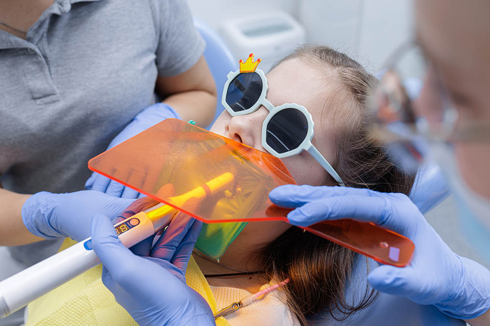 Dentist cures composite under orange shield with rubber dam while a child wears fun glasses, highlighting safe pediatric restoration and isolation in clinic.